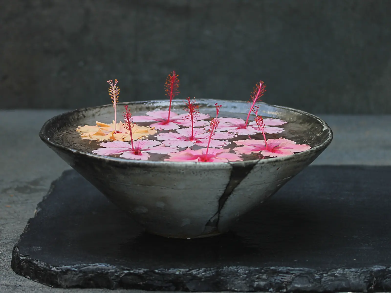 A raku bowl of water with floating pink hibiscus blooms on a slate slab